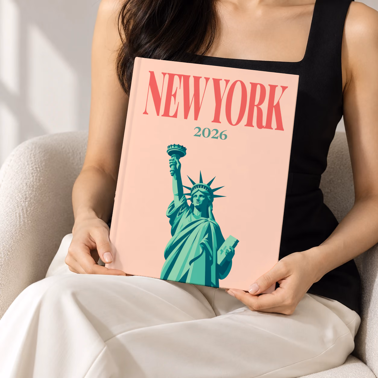 New York hardcover photo book with the illustrated cover of the Statue of Liberty on peach, held upright in two hands by a woman in a black top, sitting in a cream boucle armchair