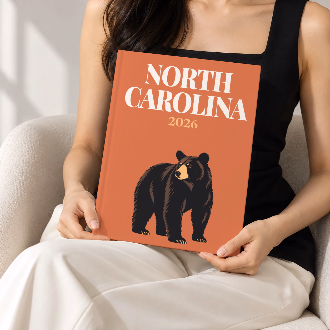 North Carolina hardcover photo book with the illustrated cover of a black bear on burnt orange, held upright in two hands by a woman in a black top, sitting in a cream boucle armchair