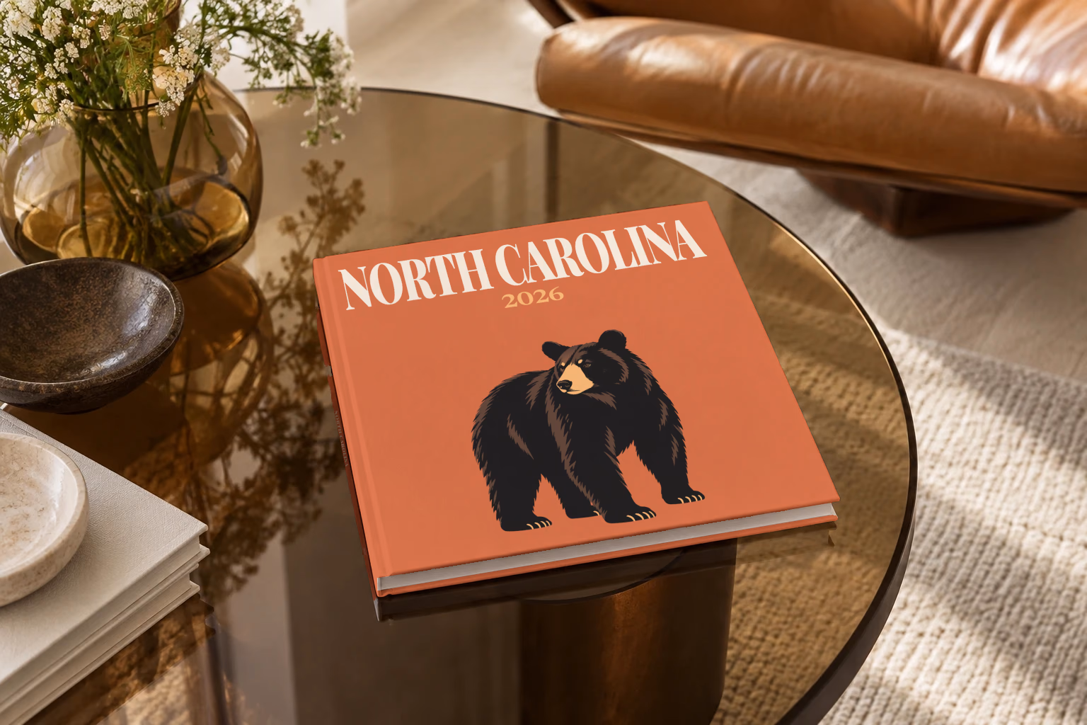 North Carolina square hardcover photo book with the illustrated cover of a black bear on burnt orange, on a smoked glass coffee table beside a leather armchair in warm afternoon light