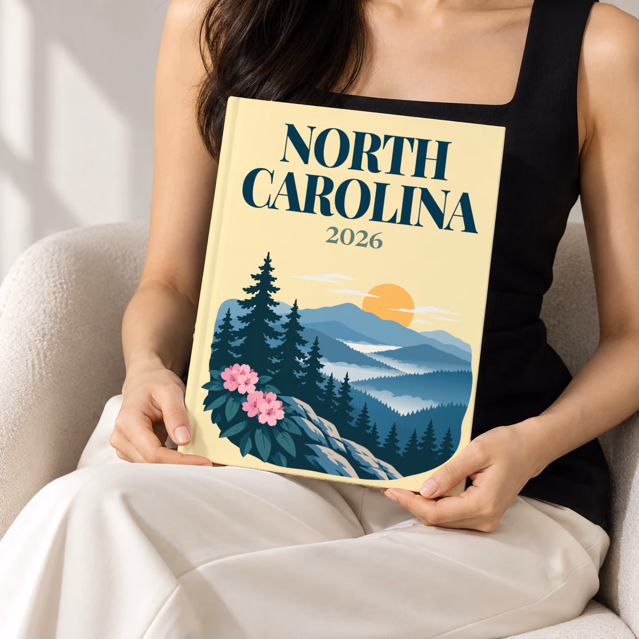 North Carolina hardcover photo book with the illustrated cover of the Blue Ridge Mountains with pine trees and pink rhododendron on pale yellow, held upright in two hands by a woman in a black top, sitting in a cream boucle armchair