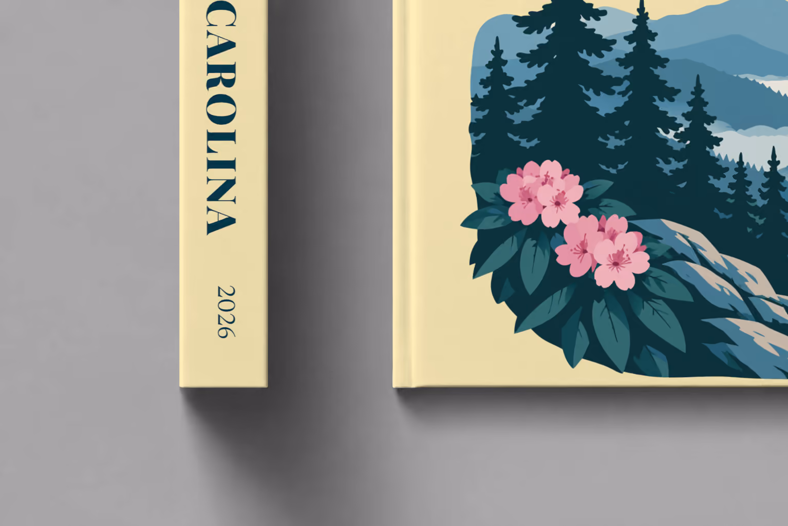 North Carolina hardcover photo book with the illustrated cover of the Blue Ridge Mountains with pine trees and pink rhododendron on pale yellow, shown close up beside its spine on a pale grey studio surface