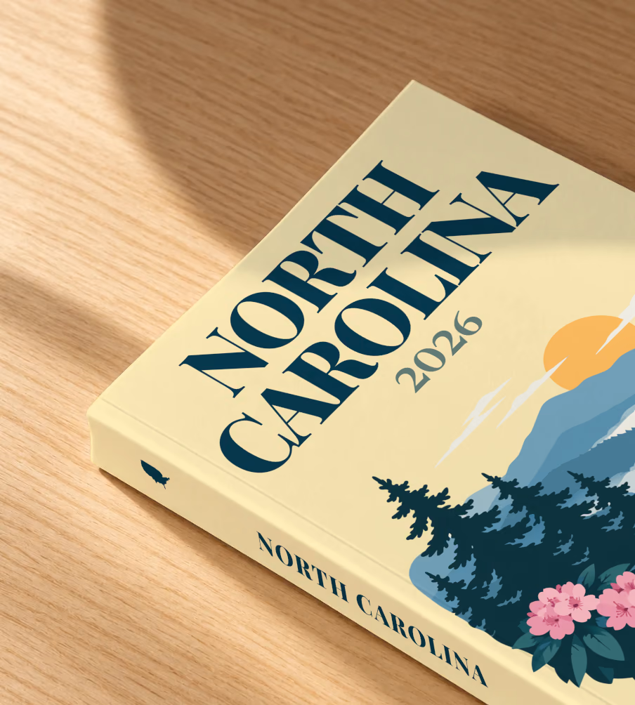 North Carolina softcover photo book with the illustrated cover of the Blue Ridge Mountains with pine trees and pink rhododendron on pale yellow, lying on an oak desk in dappled afternoon sunlight