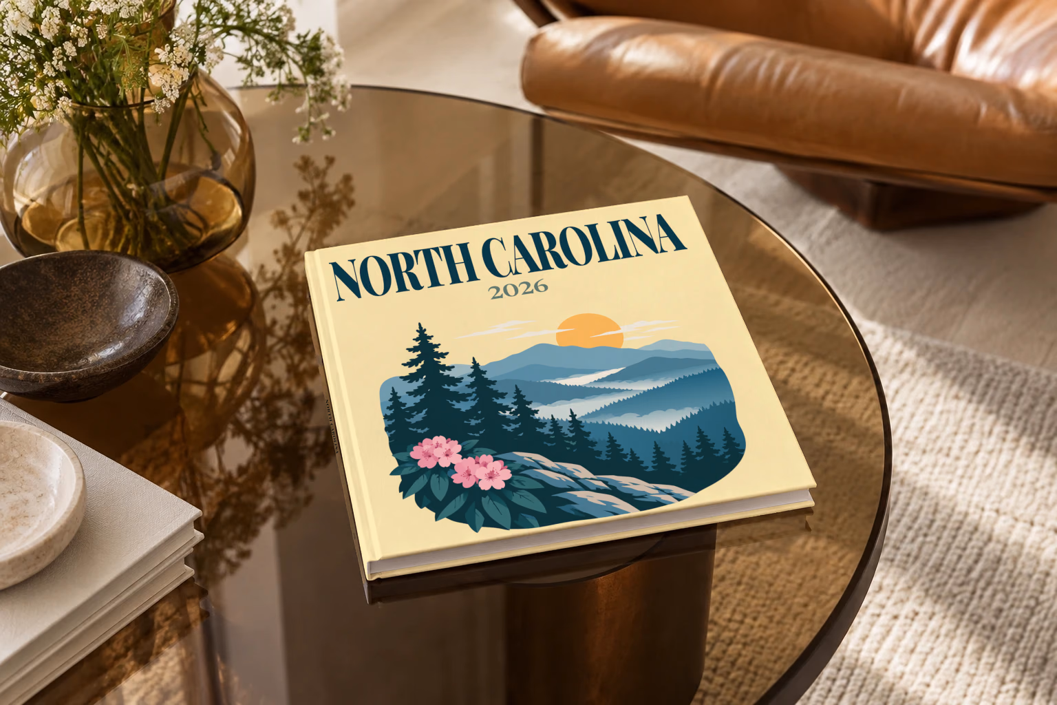 North Carolina square hardcover photo book with the illustrated cover of the Blue Ridge Mountains with pine trees and pink rhododendron on pale yellow, on a smoked glass coffee table beside a leather armchair in warm afternoon light