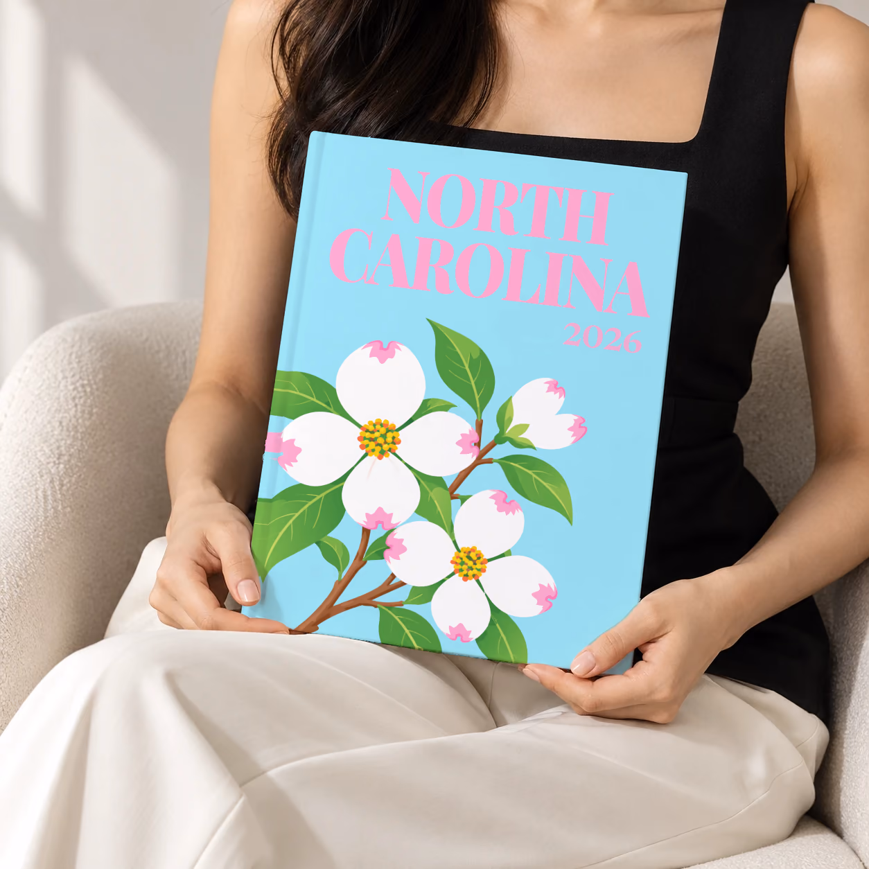 North Carolina hardcover photo book with the illustrated cover of white dogwood blossoms, the state flower, on pale aqua, held upright in two hands by a woman in a black top, sitting in a cream boucle armchair