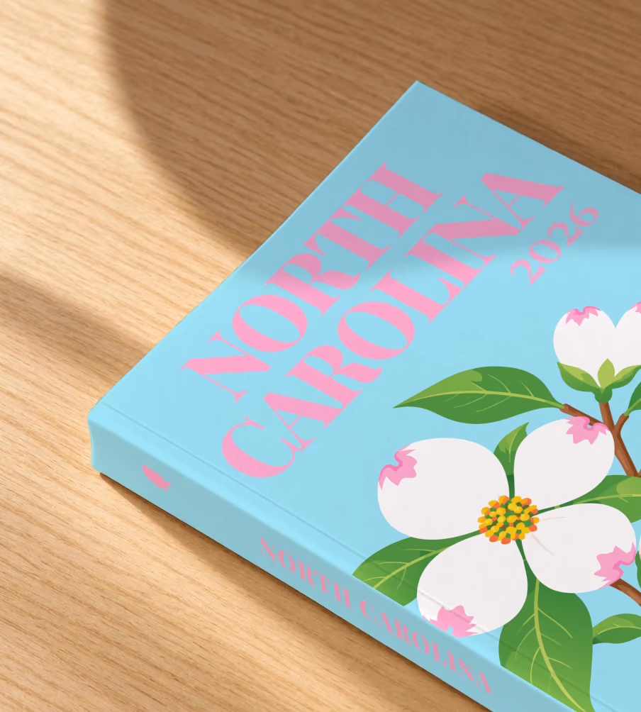 North Carolina softcover photo book with the illustrated cover of white dogwood blossoms, the state flower, on pale aqua, lying on an oak desk in dappled afternoon sunlight