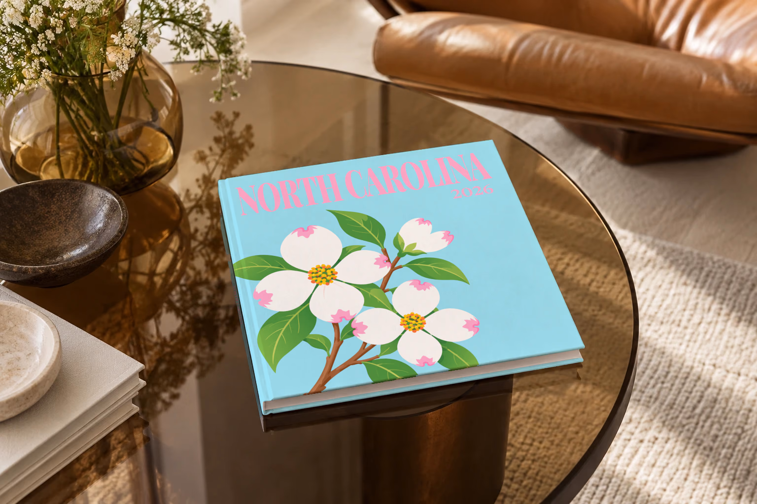 North Carolina square hardcover photo book with the illustrated cover of white dogwood blossoms, the state flower, on pale aqua, on a smoked glass coffee table beside a leather armchair in warm afternoon light
