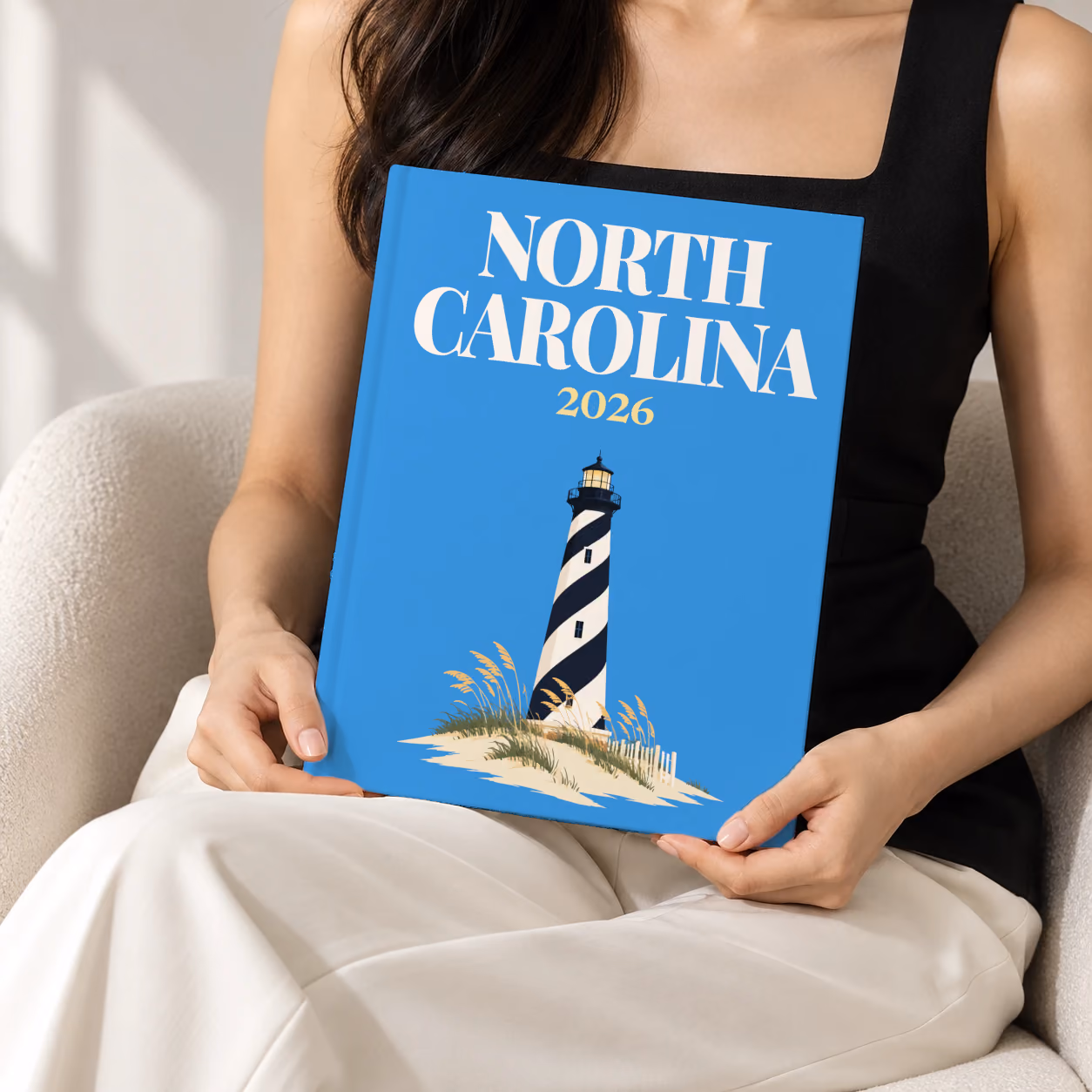 North Carolina hardcover photo book with the illustrated cover of the black and white spiral Cape Hatteras Lighthouse on the dunes on blue, held upright in two hands by a woman in a black top, sitting in a cream boucle armchair