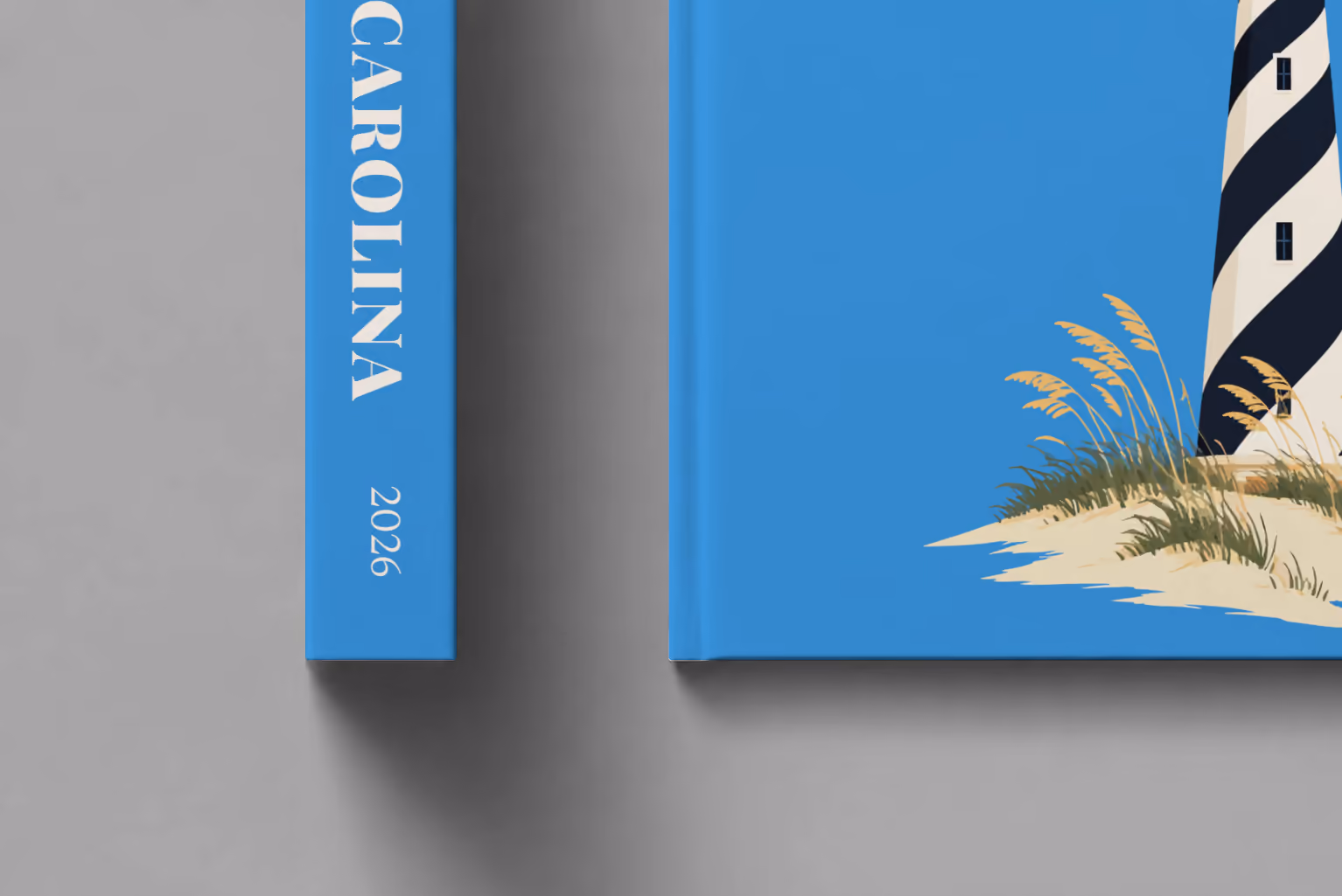 North Carolina hardcover photo book with the illustrated cover of the black and white spiral Cape Hatteras Lighthouse on the dunes on blue, shown close up beside its spine on a pale grey studio surface