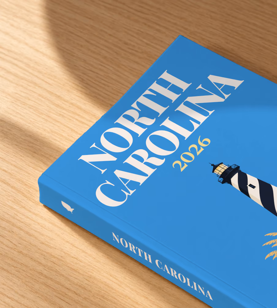 North Carolina softcover photo book with the illustrated cover of the black and white spiral Cape Hatteras Lighthouse on the dunes on blue, lying on an oak desk in dappled afternoon sunlight