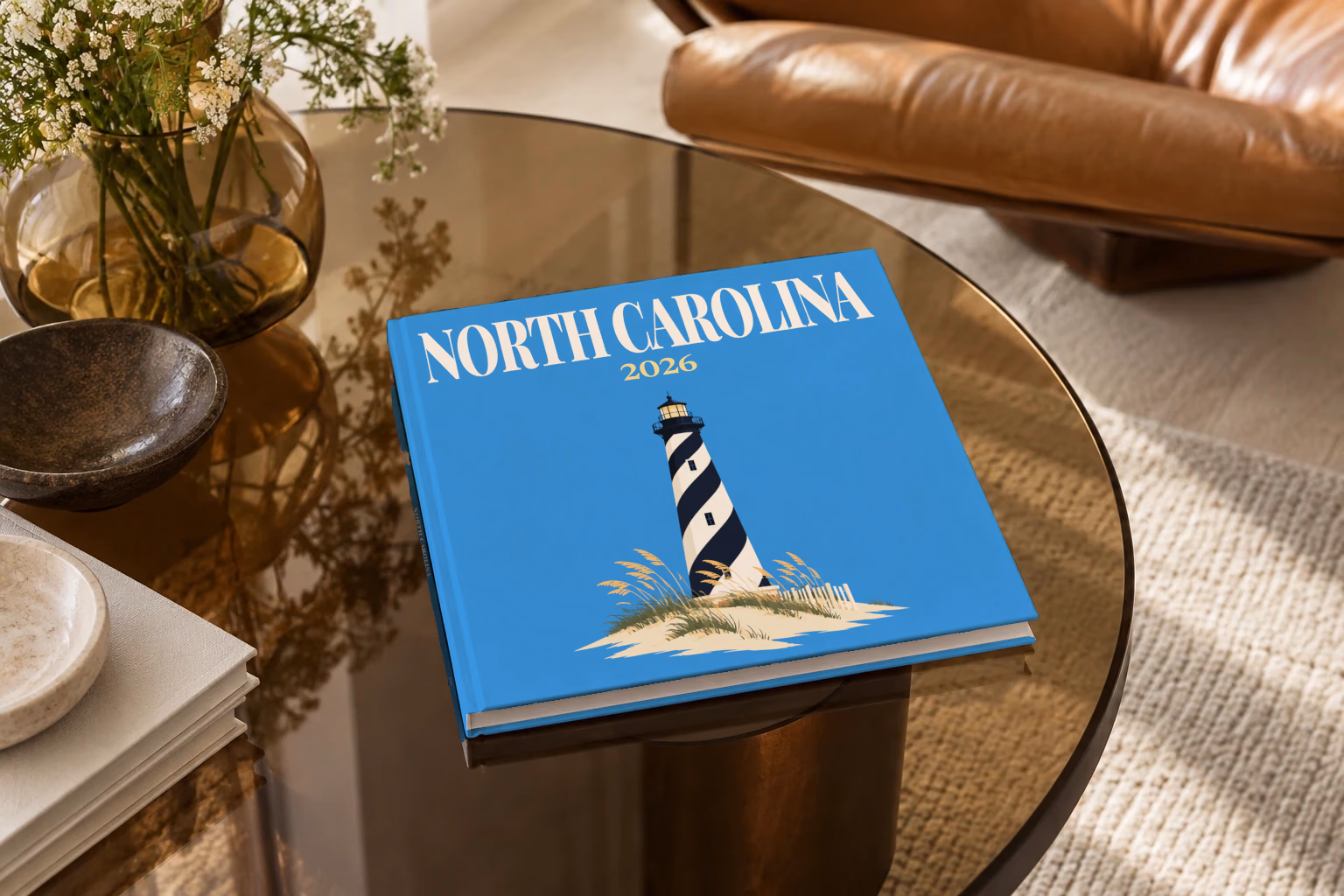 North Carolina square hardcover photo book with the illustrated cover of the black and white spiral Cape Hatteras Lighthouse on the dunes on blue, on a smoked glass coffee table beside a leather armchair in warm afternoon light
