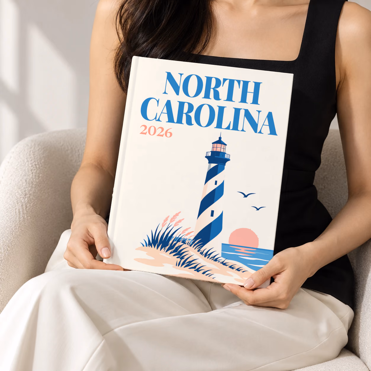 North Carolina hardcover photo book with the illustrated cover of a blue-striped lighthouse on the Outer Banks dunes at sunset on off-white, held upright in two hands by a woman in a black top, sitting in a cream boucle armchair