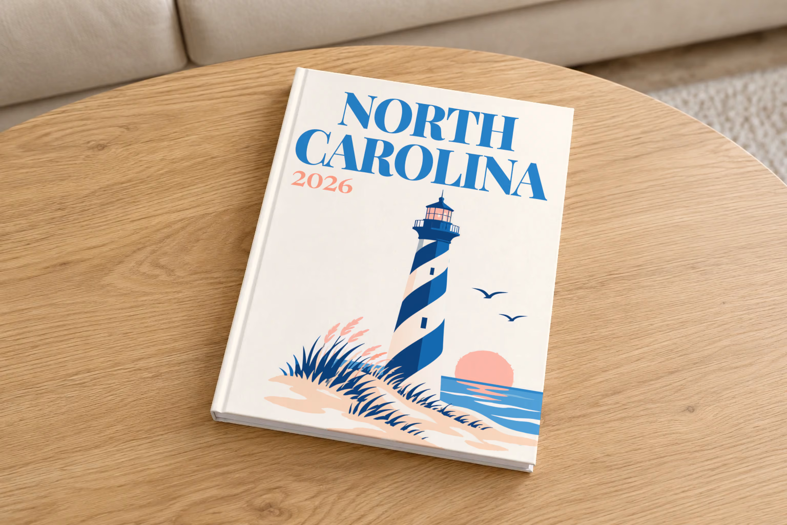 North Carolina hardcover photo book with the illustrated cover of a blue-striped lighthouse on the Outer Banks dunes at sunset on off-white, on a round oak coffee table in a bright living room