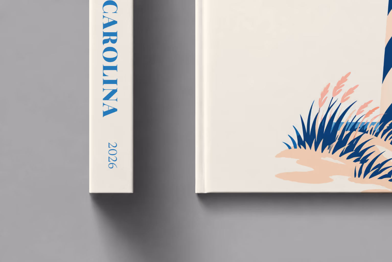 North Carolina hardcover photo book with the illustrated cover of a blue-striped lighthouse on the Outer Banks dunes at sunset on off-white, shown close up beside its spine on a pale grey studio surface