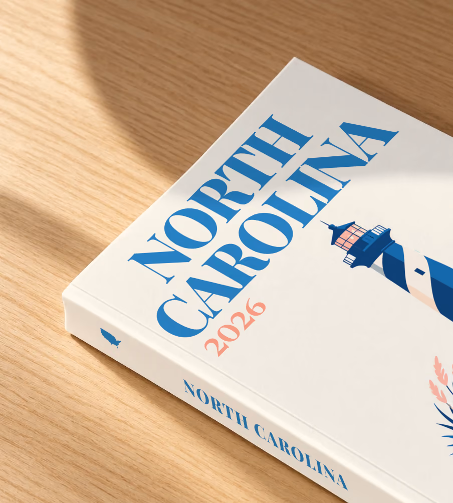 North Carolina softcover photo book with the illustrated cover of a blue-striped lighthouse on the Outer Banks dunes at sunset on off-white, lying on an oak desk in dappled afternoon sunlight