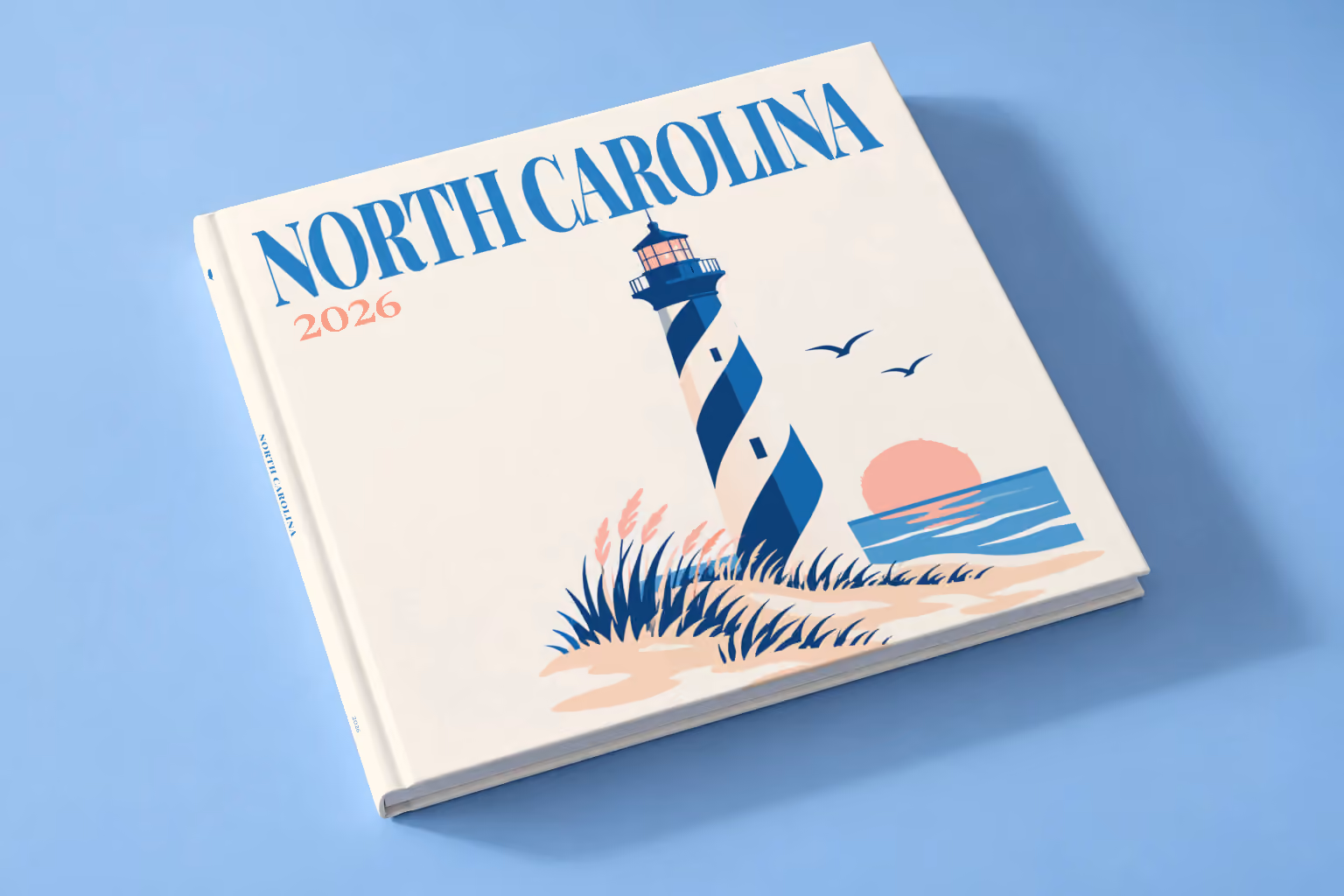North Carolina square hardcover photo book with the illustrated cover of a blue-striped lighthouse on the Outer Banks dunes at sunset on off-white, lying on a pale blue studio backdrop
