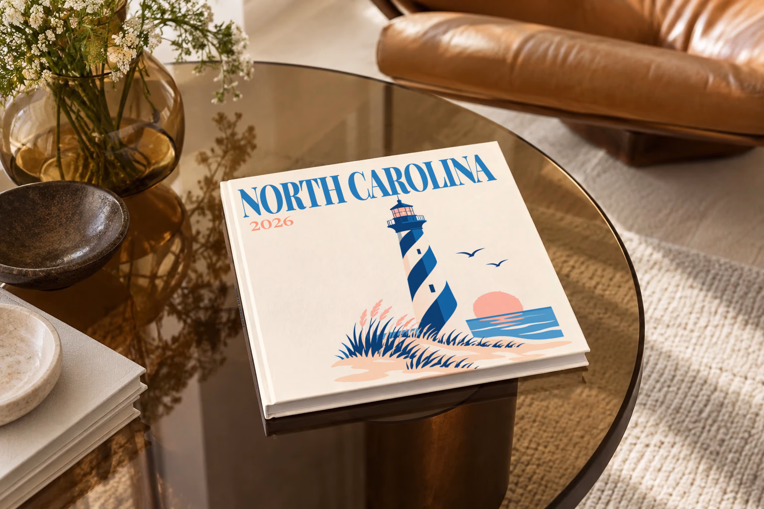 North Carolina square hardcover photo book with the illustrated cover of a blue-striped lighthouse on the Outer Banks dunes at sunset on off-white, on a smoked glass coffee table beside a leather armchair in warm afternoon light