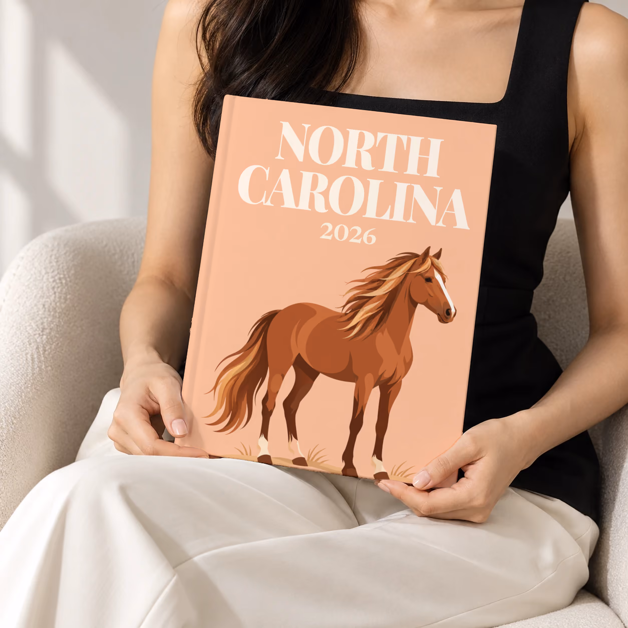 North Carolina hardcover photo book with the illustrated cover of a wild Outer Banks horse on peach, held upright in two hands by a woman in a black top, sitting in a cream boucle armchair