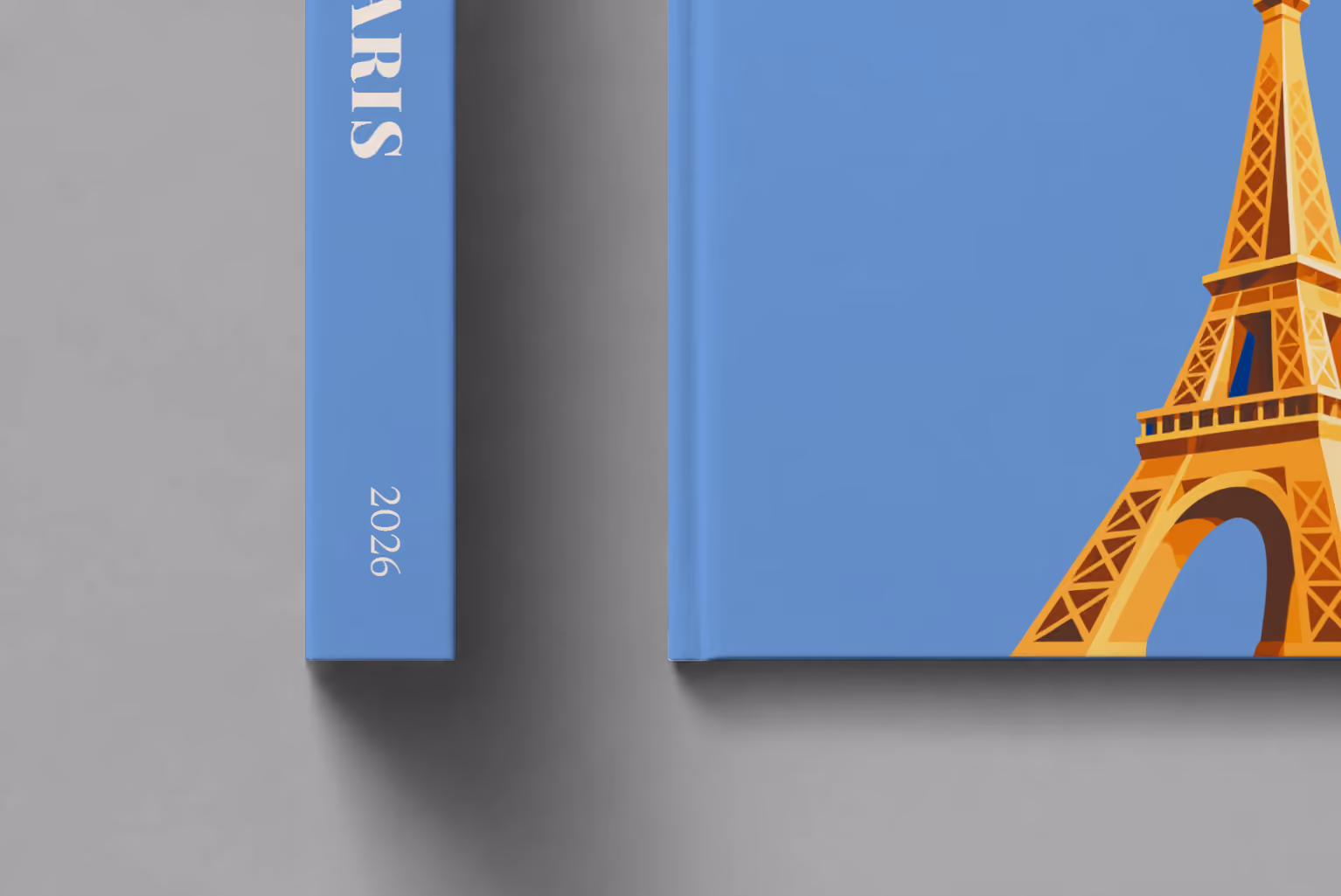 Paris hardcover photo book with the illustrated cover of a gold Eiffel Tower on blue, shown close up beside its spine on a pale grey studio surface