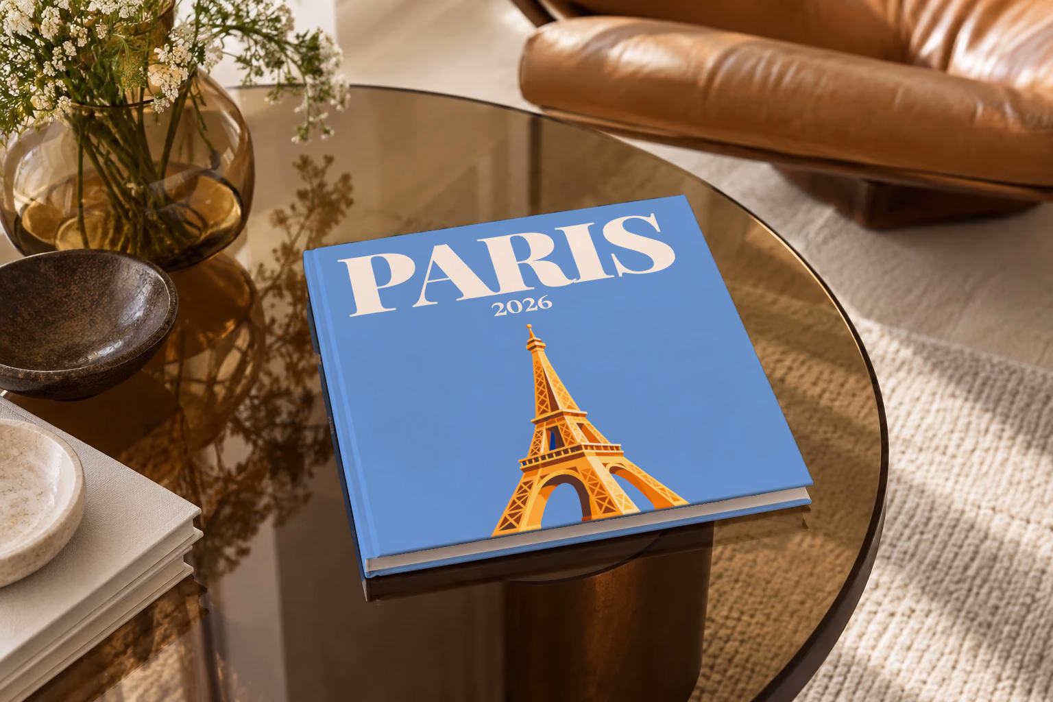 Paris square hardcover photo book with the illustrated cover of a gold Eiffel Tower on blue, on a smoked glass coffee table beside a leather armchair in warm afternoon light