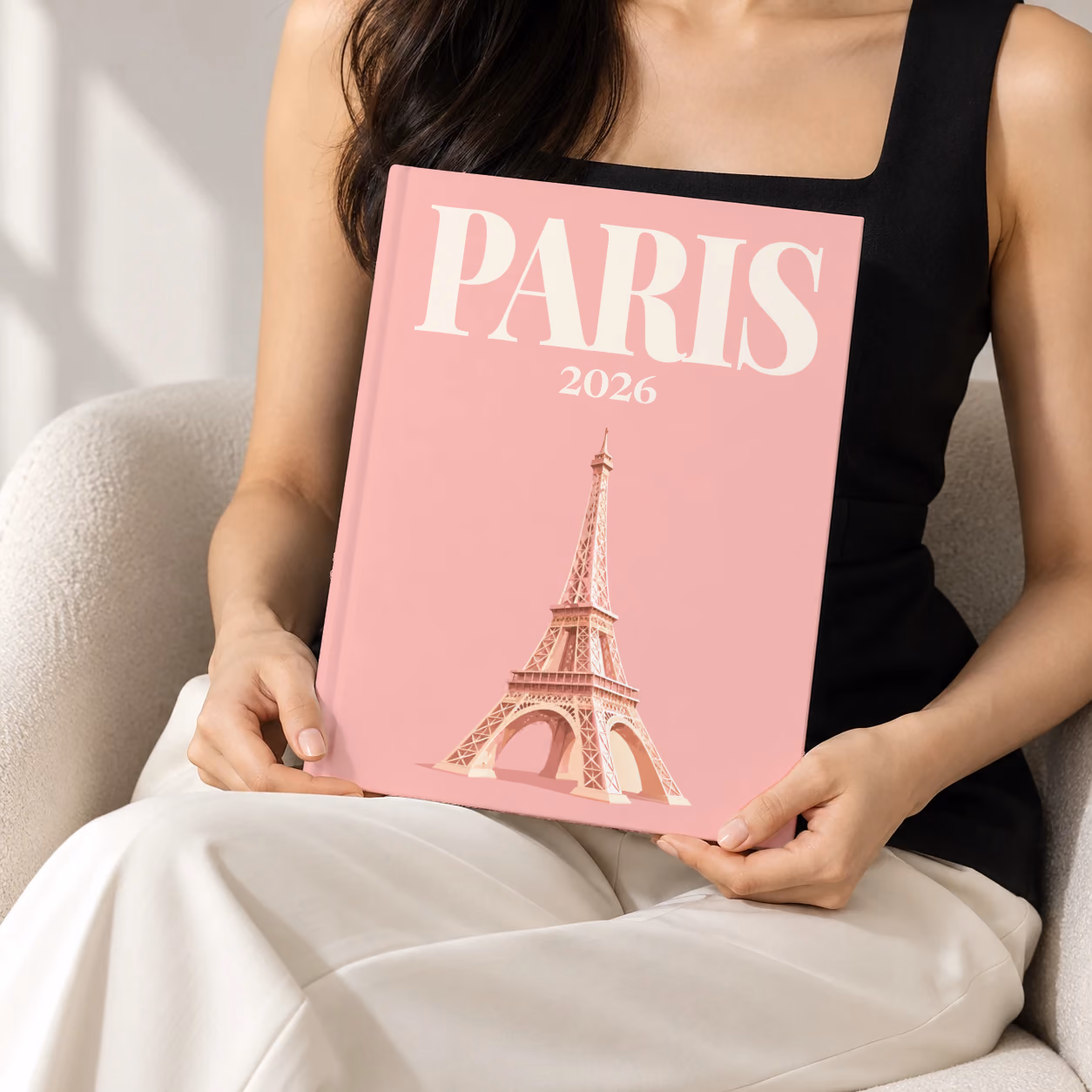 Paris hardcover photo book with the illustrated cover of the Eiffel Tower on blush pink, held upright in two hands by a woman in a black top, sitting in a cream boucle armchair