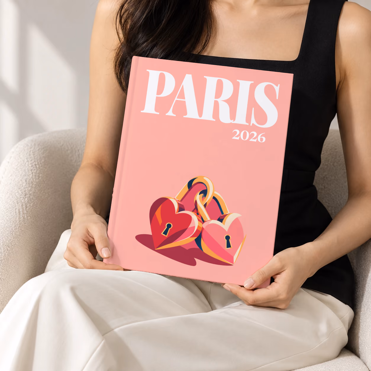Paris hardcover photo book with the illustrated cover of heart-shaped love locks on salmon pink, held upright in two hands by a woman in a black top, sitting in a cream boucle armchair