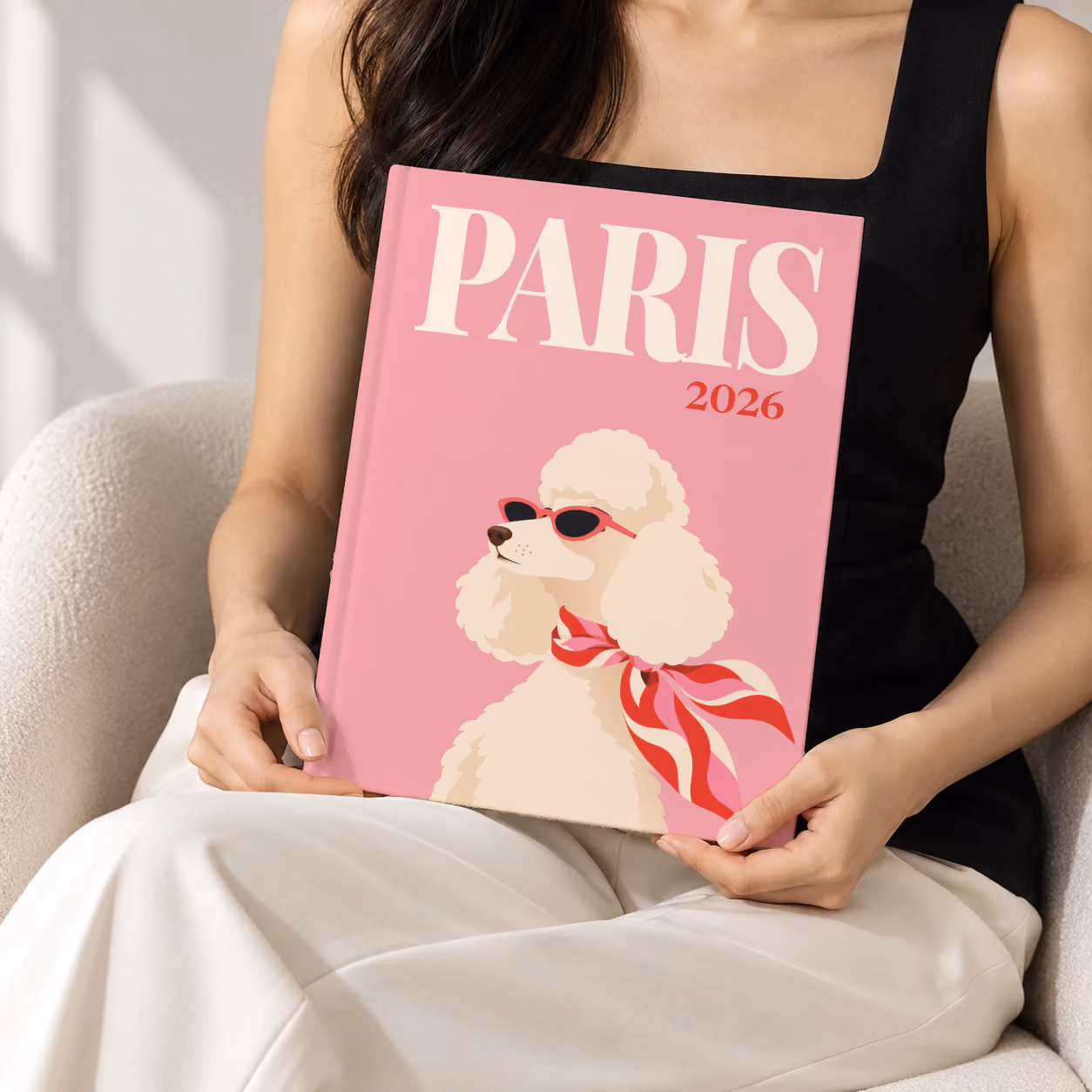 Paris hardcover photo book with the illustrated cover of a poodle in sunglasses and a scarf on pink, held upright in two hands by a woman in a black top, sitting in a cream boucle armchair