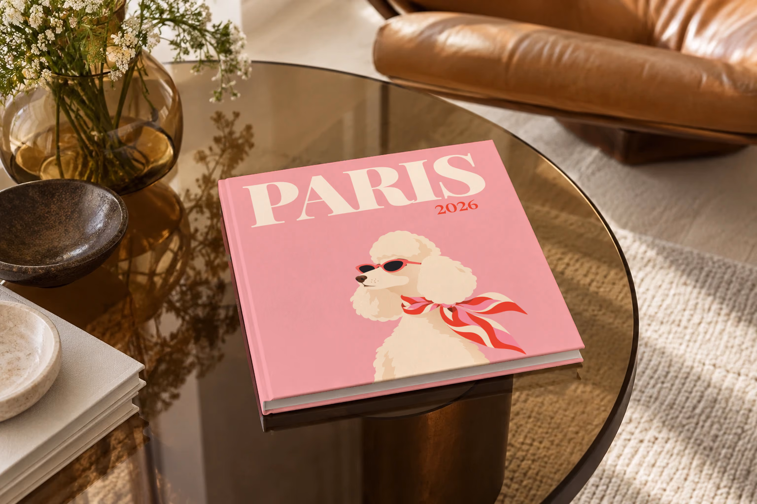 Paris square hardcover photo book with the illustrated cover of a poodle in sunglasses and a scarf on pink, on a smoked glass coffee table beside a leather armchair in warm afternoon light