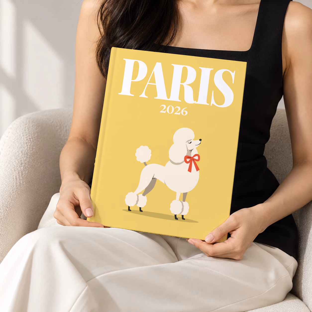Paris hardcover photo book with the illustrated cover of a white poodle with a red bow on yellow, held upright in two hands by a woman in a black top, sitting in a cream boucle armchair