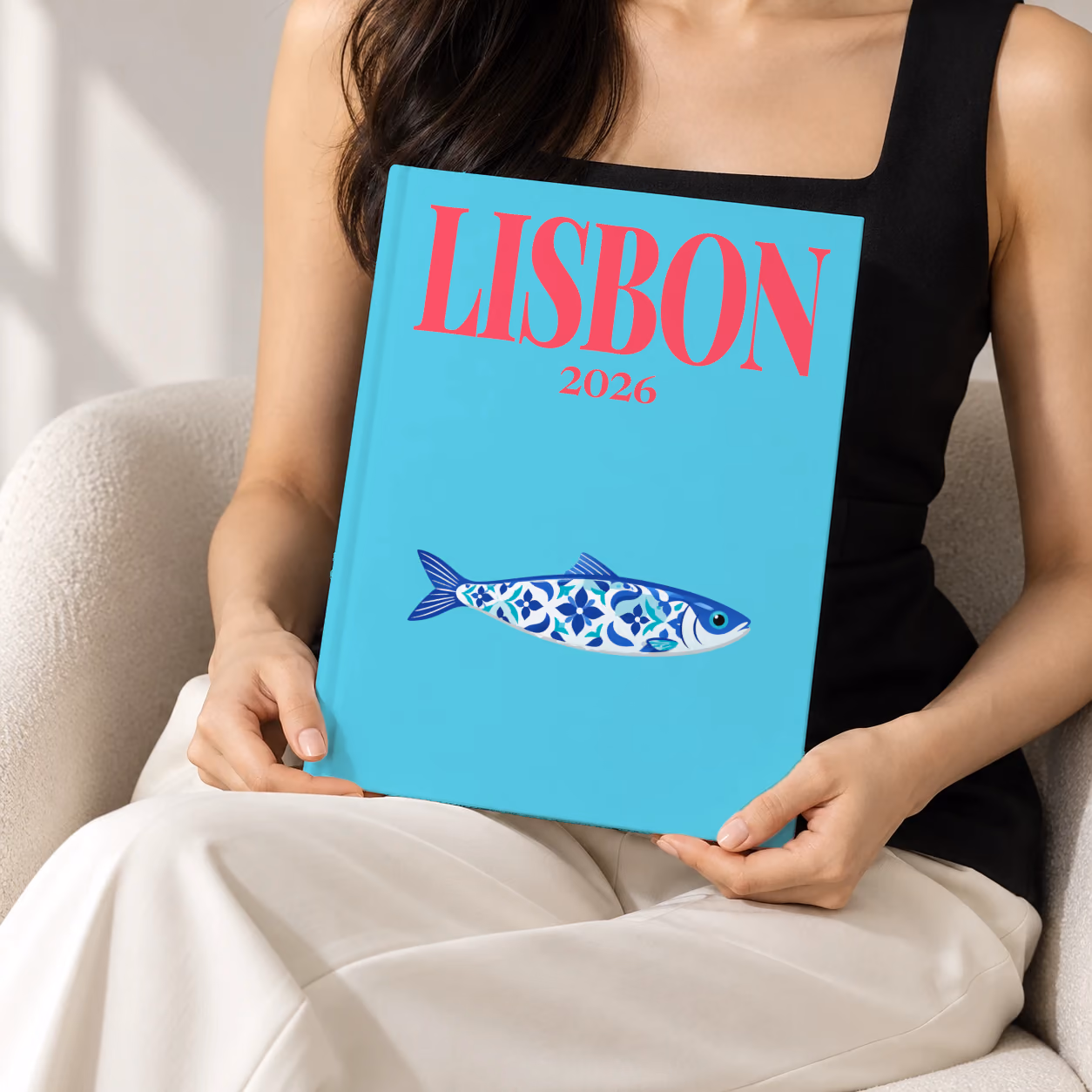 Portugal hardcover photo book with the Lisbon illustrated cover of a blue azulejo-tiled Portuguese sardine on turquoise, held upright in two hands by a woman in a black top, sitting in a cream boucle armchair
