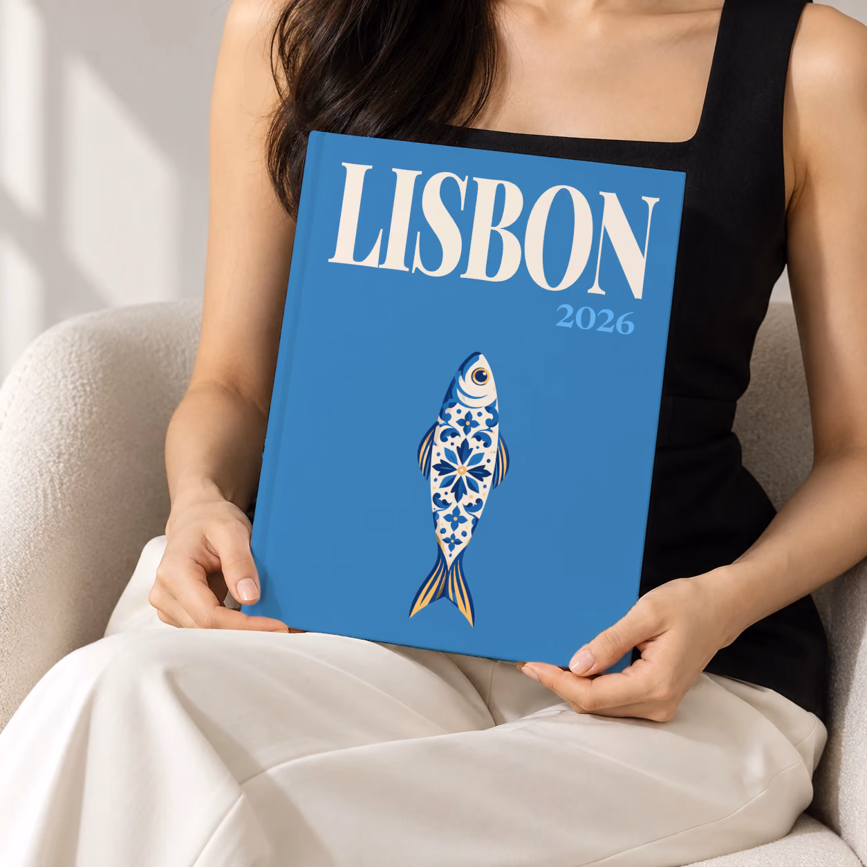 Portugal hardcover photo book with the Lisbon illustrated cover of a blue and white azulejo-tiled sardine on denim blue, held upright in two hands by a woman in a black top, sitting in a cream boucle armchair