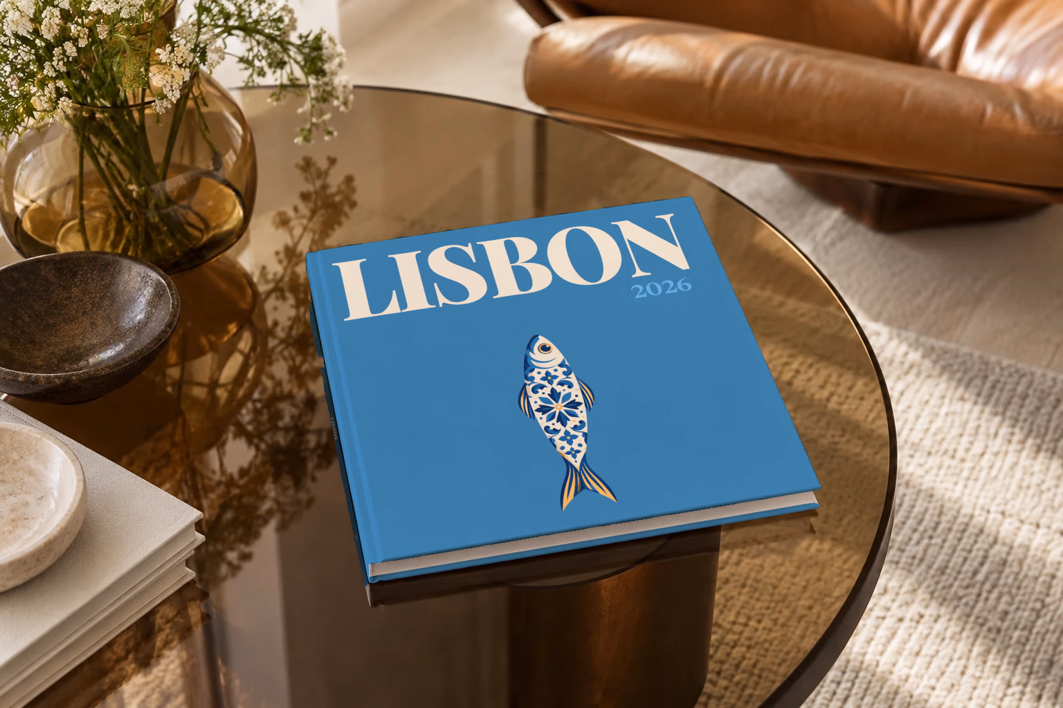 Portugal square hardcover photo book with the Lisbon illustrated cover of a blue and white azulejo-tiled sardine on denim blue, on a smoked glass coffee table beside a leather armchair in warm afternoon light