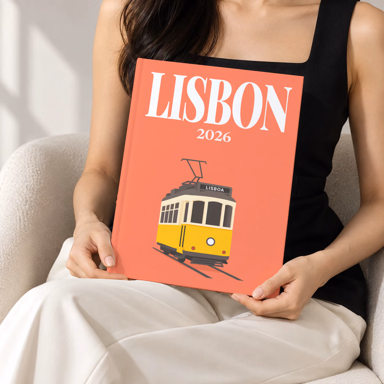 Portugal hardcover photo book with the Lisbon illustrated cover of a yellow vintage Lisbon Tram 28 on coral, held upright in two hands by a woman in a black top, sitting in a cream boucle armchair