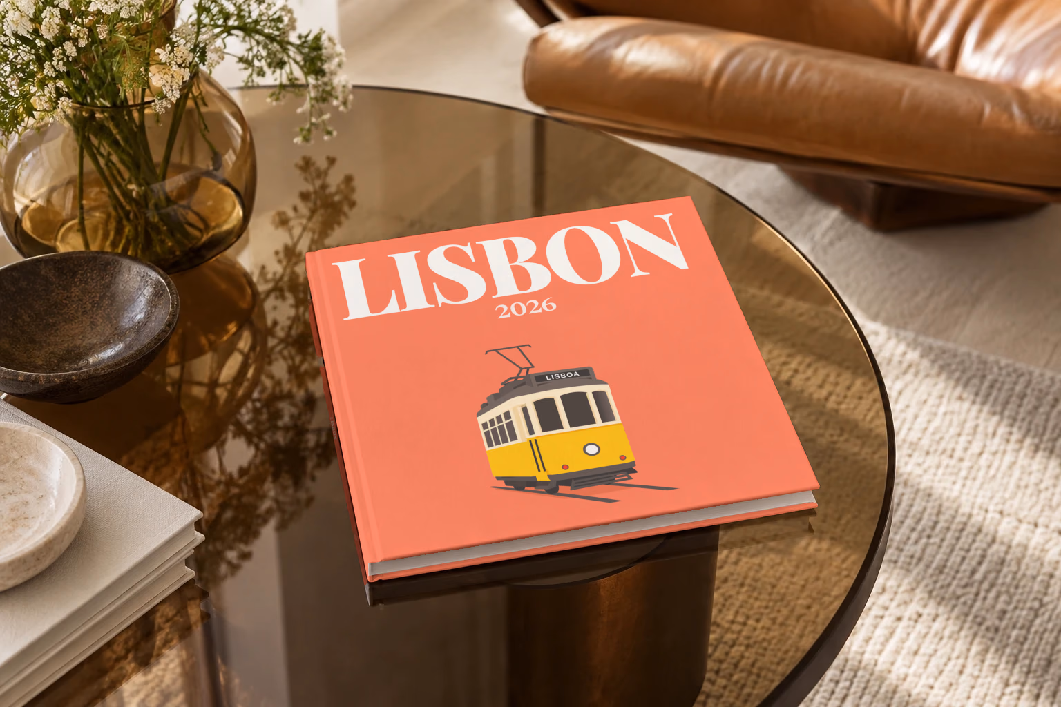 Portugal square hardcover photo book with the Lisbon illustrated cover of a yellow vintage Lisbon Tram 28 on coral, on a smoked glass coffee table beside a leather armchair in warm afternoon light