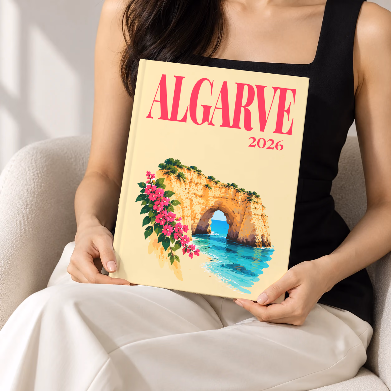Portugal hardcover photo book with the Algarve illustrated cover of a golden Algarve sea arch over turquoise water with pink bougainvillea on pale yellow, held upright in two hands by a woman in a black top, sitting in a cream boucle armchair