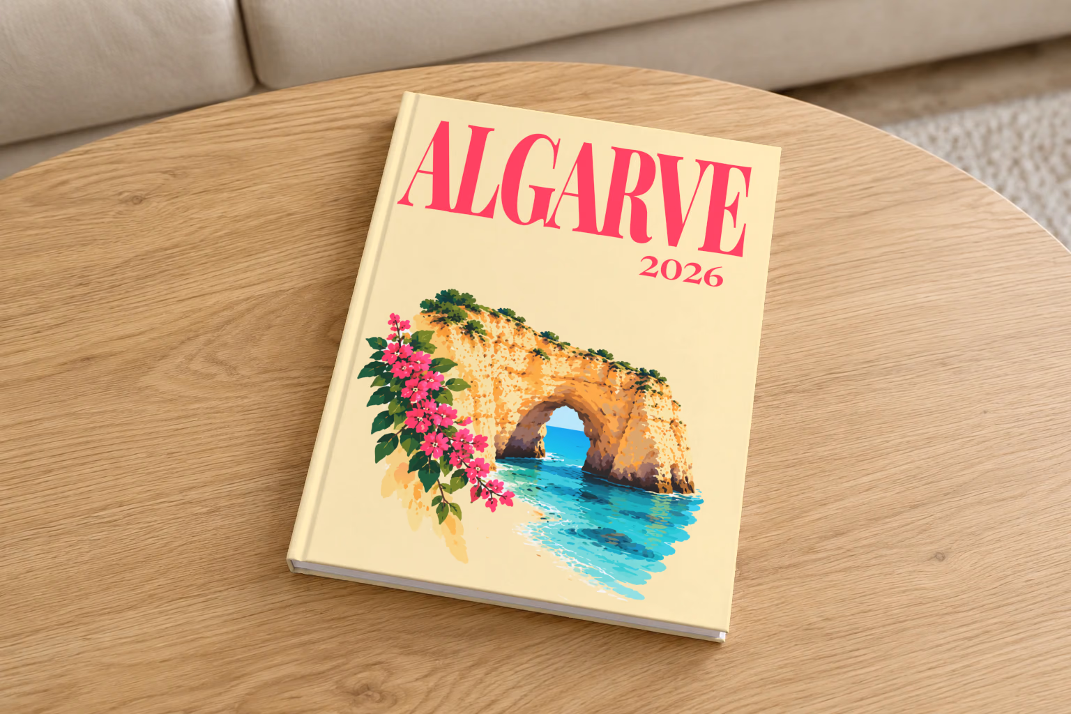 Portugal hardcover photo book with the Algarve illustrated cover of a golden Algarve sea arch over turquoise water with pink bougainvillea on pale yellow, on a round oak coffee table in a bright living room