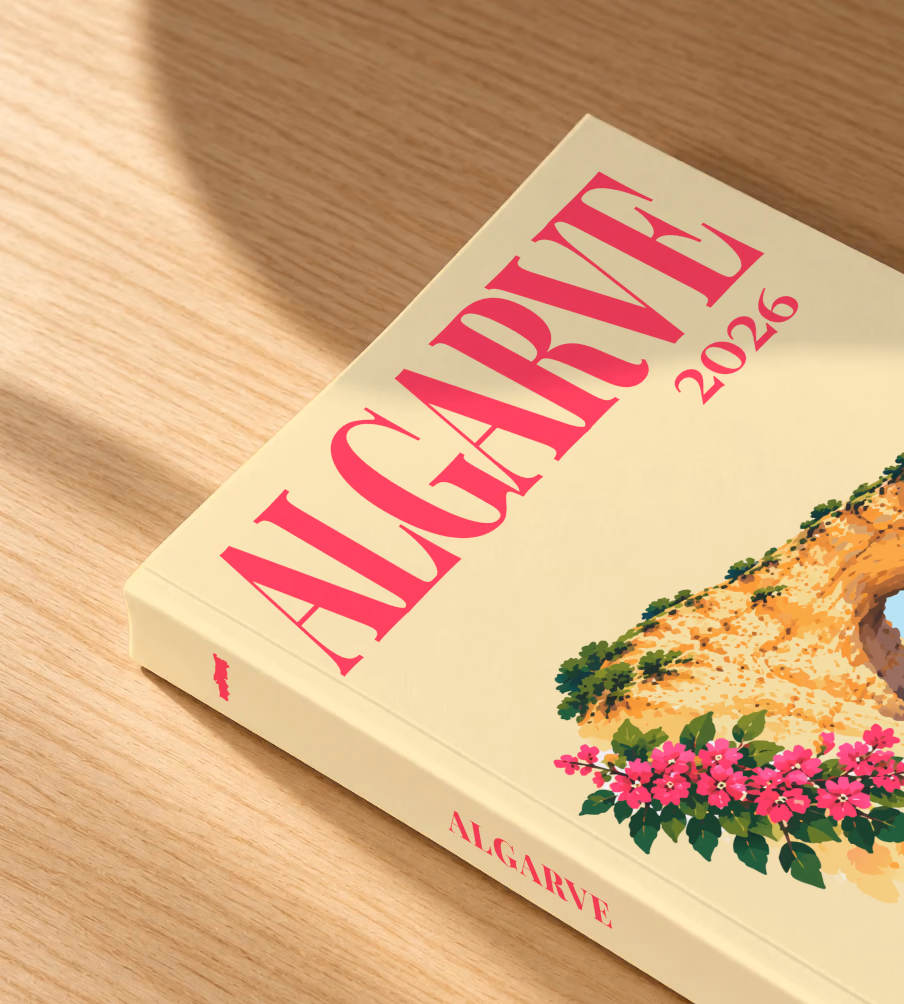 Portugal softcover photo book with the Algarve illustrated cover of a golden Algarve sea arch over turquoise water with pink bougainvillea on pale yellow, lying on an oak desk in dappled afternoon sunlight