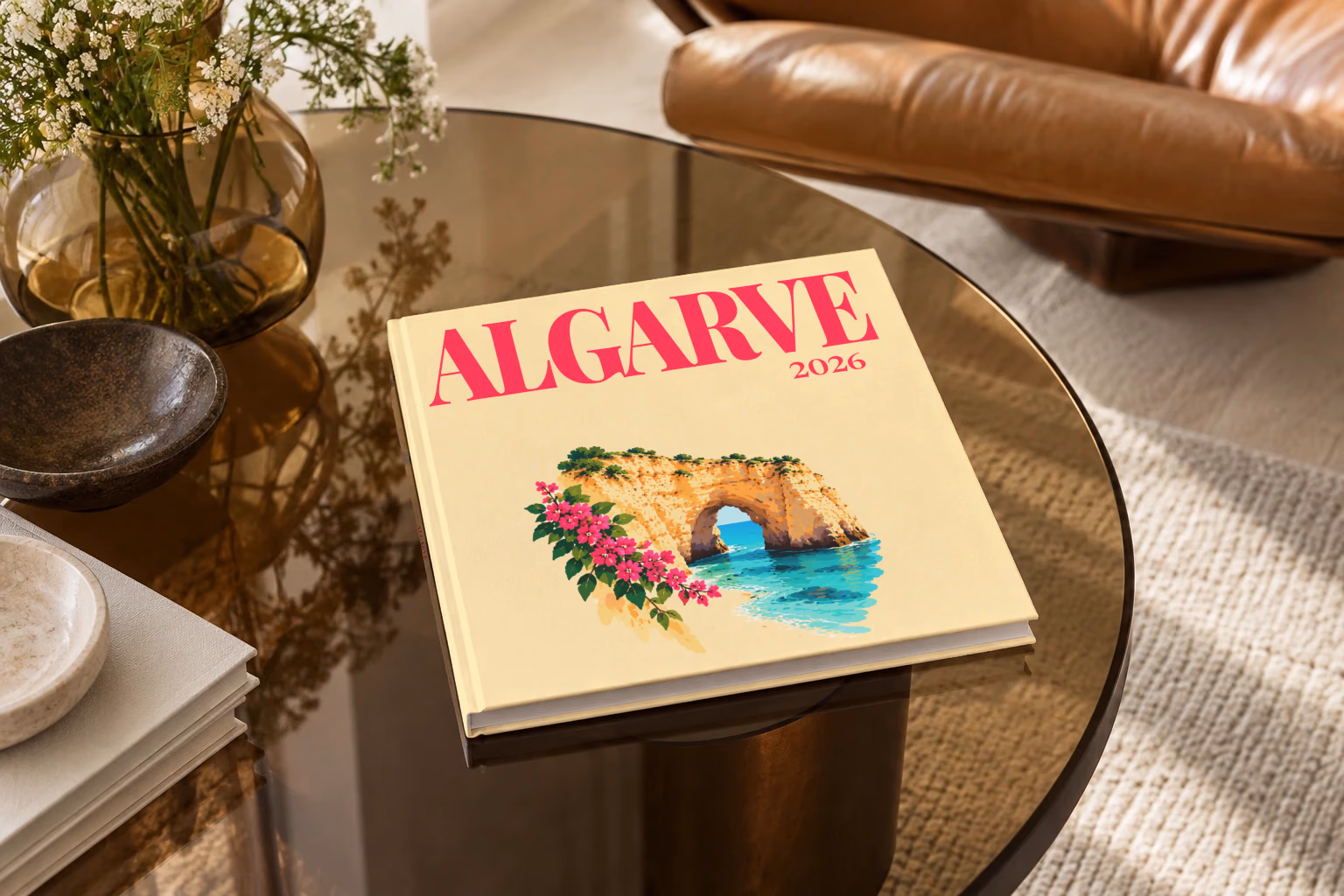 Portugal square hardcover photo book with the Algarve illustrated cover of a golden Algarve sea arch over turquoise water with pink bougainvillea on pale yellow, on a smoked glass coffee table beside a leather armchair in warm afternoon light