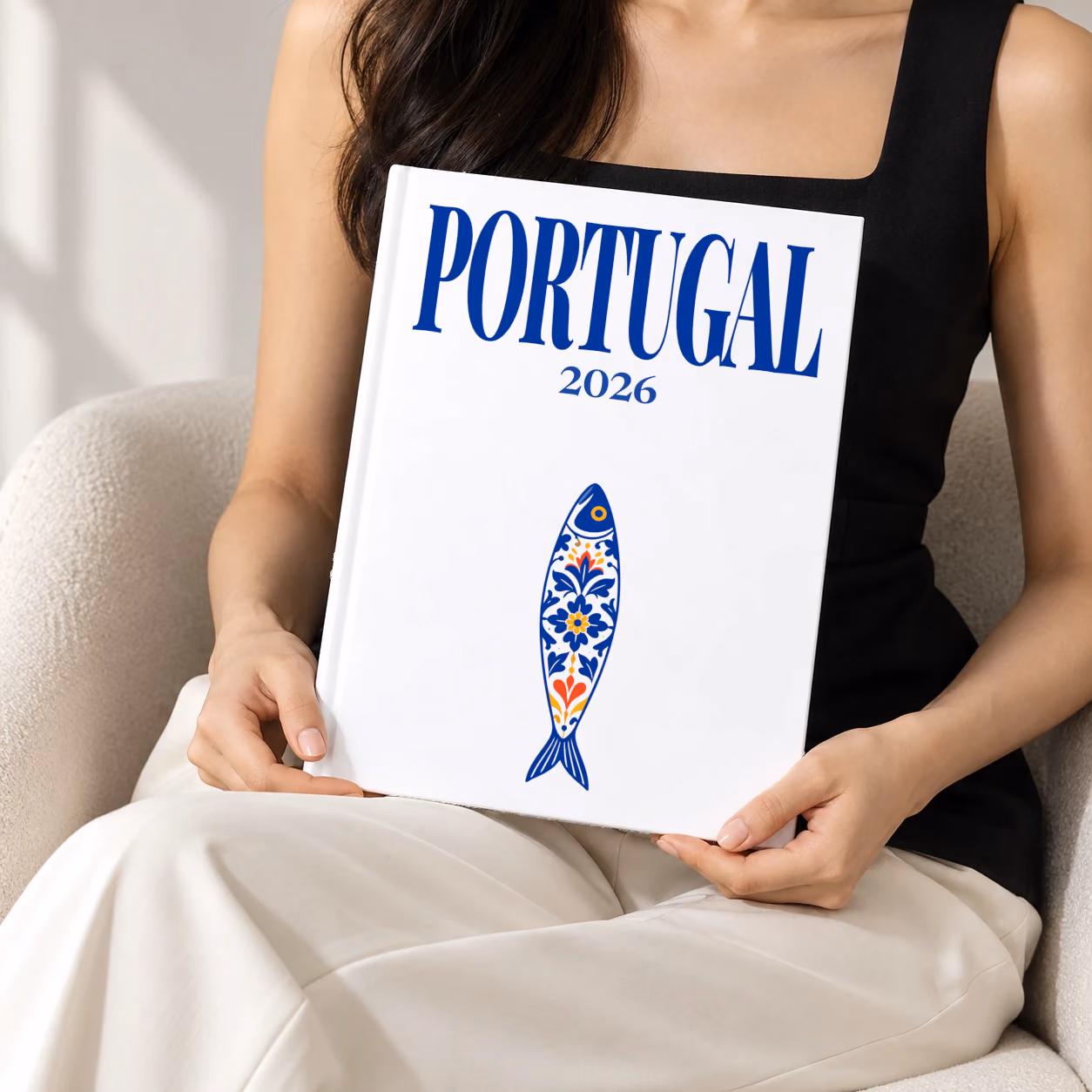Portugal hardcover photo book with the illustrated cover of a blue and white azulejo-tiled Portuguese sardine on white, held upright in two hands by a woman in a black top, sitting in a cream boucle armchair