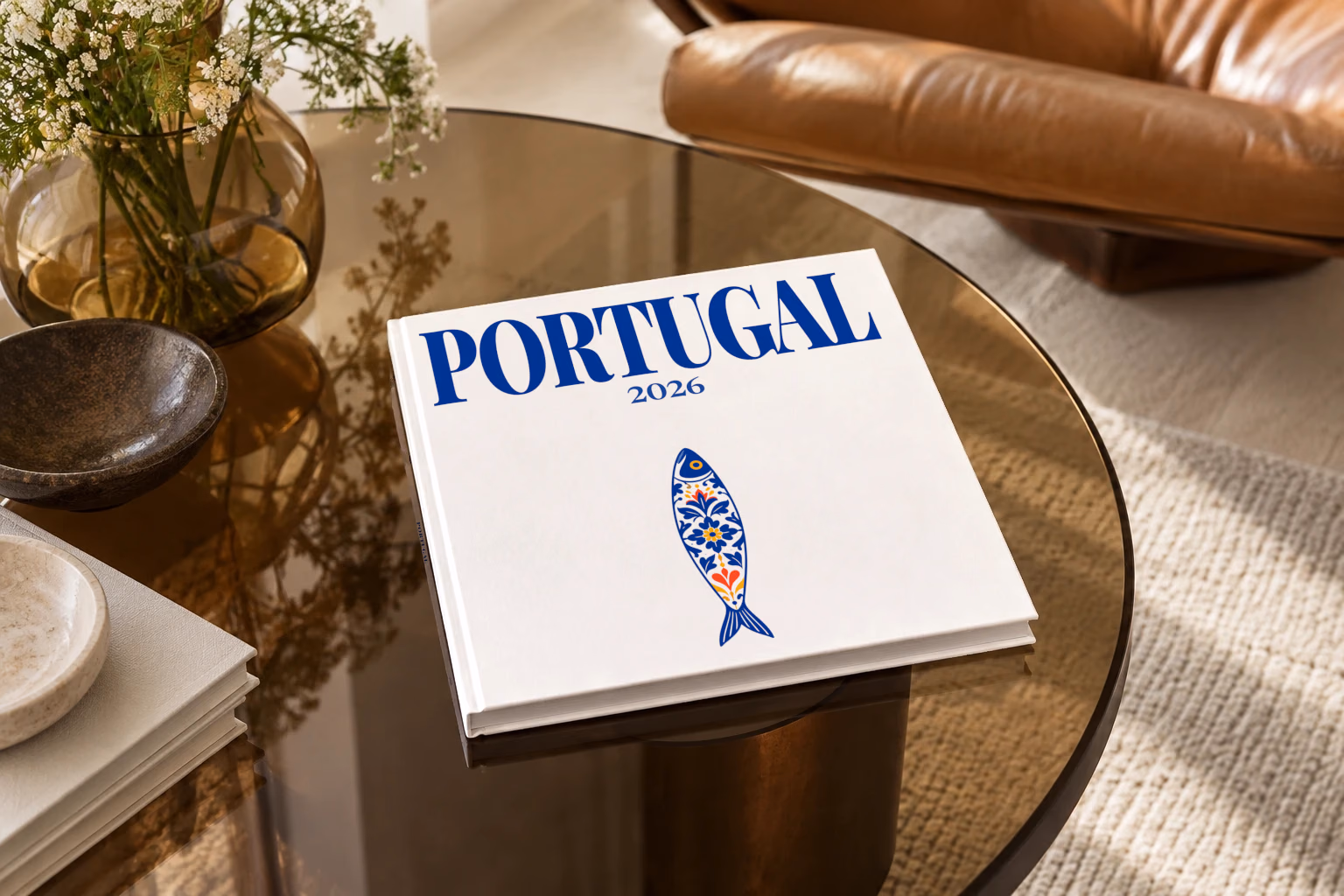 Portugal square hardcover photo book with the illustrated cover of a blue and white azulejo-tiled Portuguese sardine on white, on a smoked glass coffee table beside a leather armchair in warm afternoon light