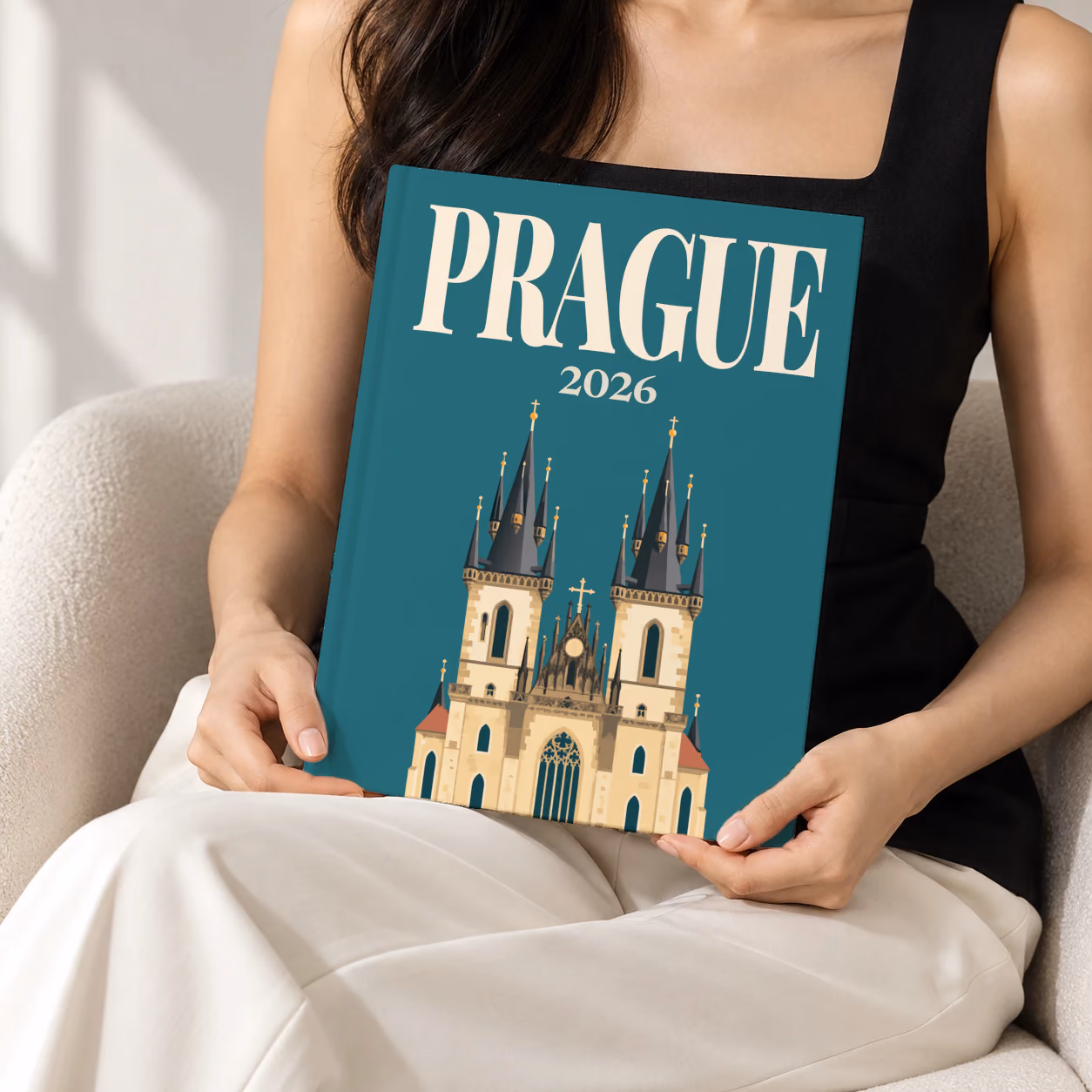 Prague hardcover photo book with the illustrated cover of the Gothic spires of the Church of Our Lady before Týn on teal, held upright in two hands by a woman in a black top, sitting in a cream boucle armchair