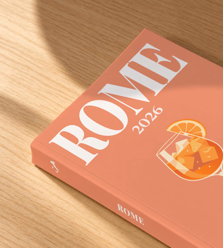 Rome softcover photo book with the illustrated cover of an Aperol spritz on coral, lying on an oak desk in dappled afternoon sunlight