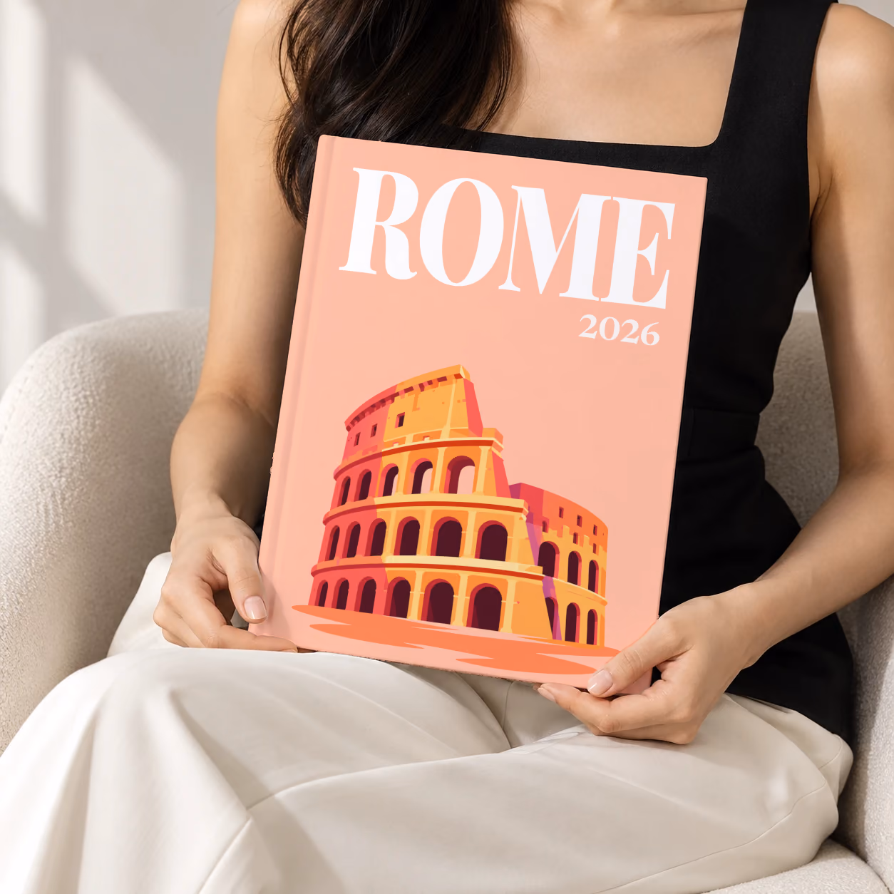 Rome hardcover photo book with the illustrated cover of the Colosseum on peach, held upright in two hands by a woman in a black top, sitting in a cream boucle armchair
