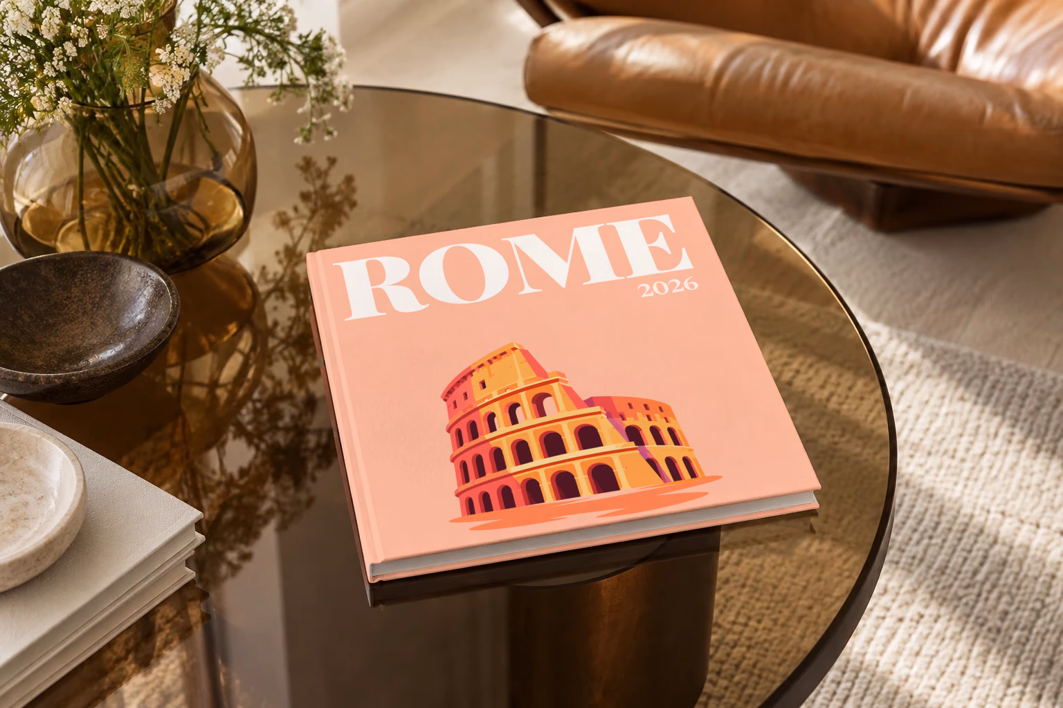 Rome square hardcover photo book with the illustrated cover of the Colosseum on peach, on a smoked glass coffee table beside a leather armchair in warm afternoon light