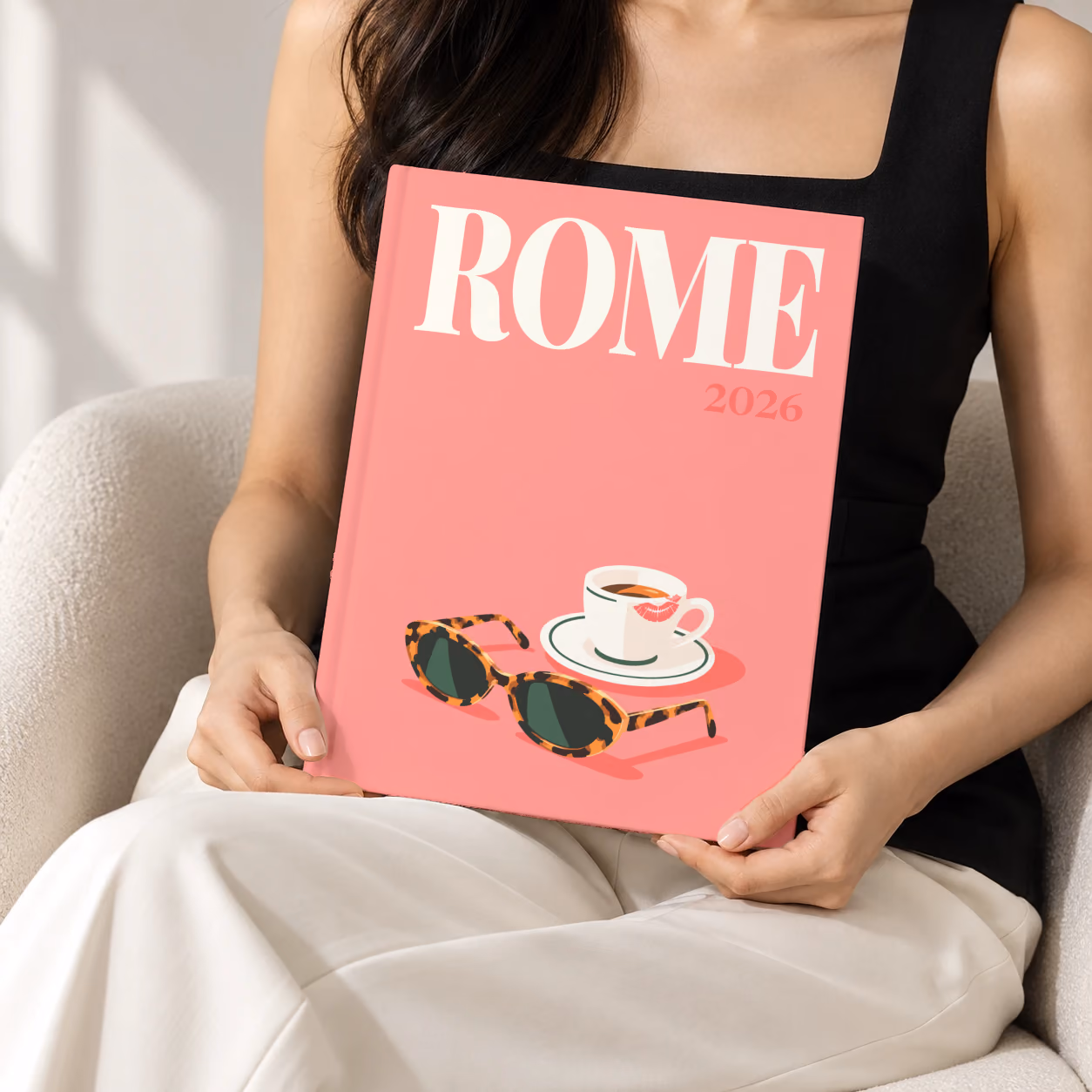 Rome hardcover photo book with the illustrated cover of an espresso cup and tortoiseshell sunglasses on salmon pink, held upright in two hands by a woman in a black top, sitting in a cream boucle armchair