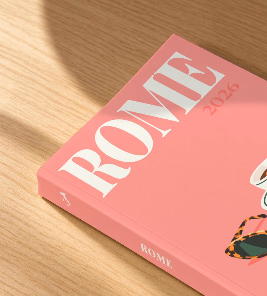 Rome softcover photo book with the illustrated cover of an espresso cup and tortoiseshell sunglasses on salmon pink, lying on an oak desk in dappled afternoon sunlight