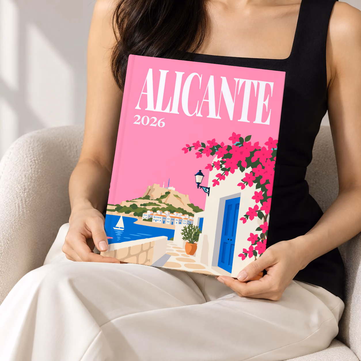 Spain hardcover photo book with the Alicante illustrated cover of a whitewashed street with bougainvillea above the sea on pink, held upright in two hands by a woman in a black top, sitting in a cream boucle armchair