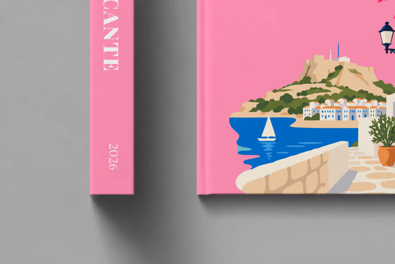 Spain hardcover photo book with the Alicante illustrated cover of a whitewashed street with bougainvillea above the sea on pink, shown close up beside its spine on a pale grey studio surface