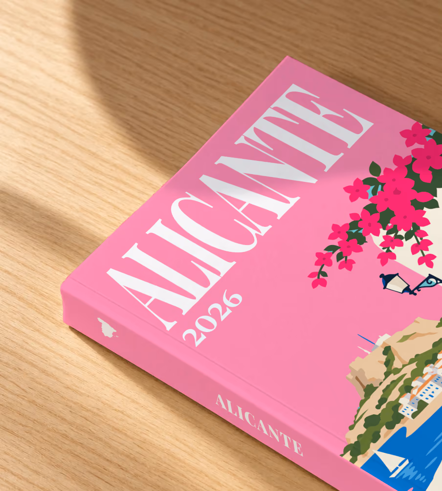 Spain softcover photo book with the Alicante illustrated cover of a whitewashed street with bougainvillea above the sea on pink, lying on an oak desk in dappled afternoon sunlight
