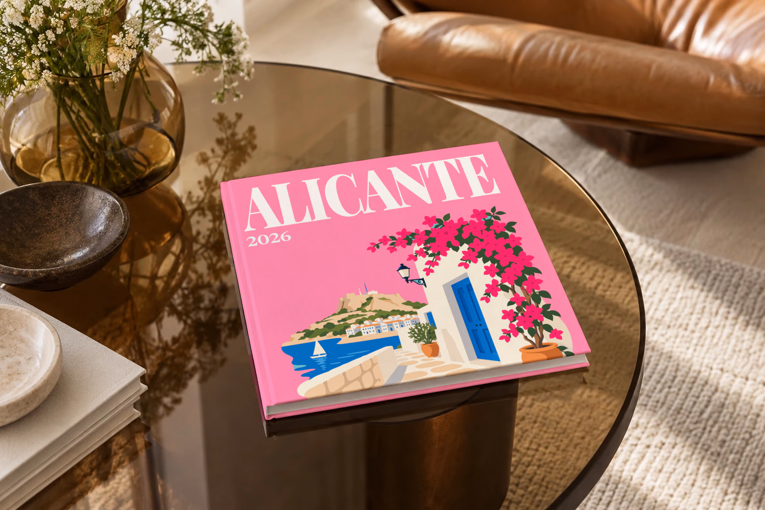 Spain square hardcover photo book with the Alicante illustrated cover of a whitewashed street with bougainvillea above the sea on pink, on a smoked glass coffee table beside a leather armchair in warm afternoon light