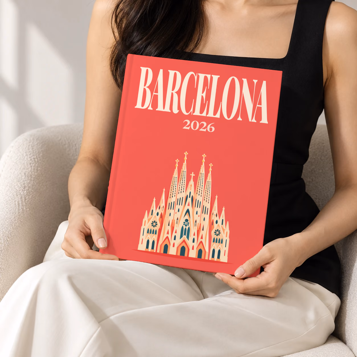 Spain hardcover photo book with the Barcelona illustrated cover of the Sagrada Família on coral red, held upright in two hands by a woman in a black top, sitting in a cream boucle armchair